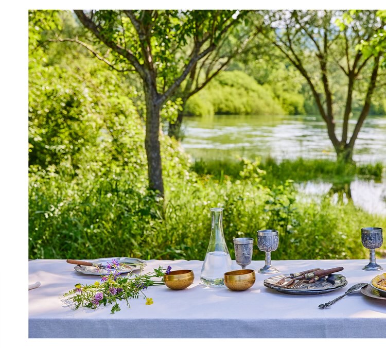 Outdoor dining table set with antique dishware, gold bowls, and flowers, next to a lake and trees on a sunny day.