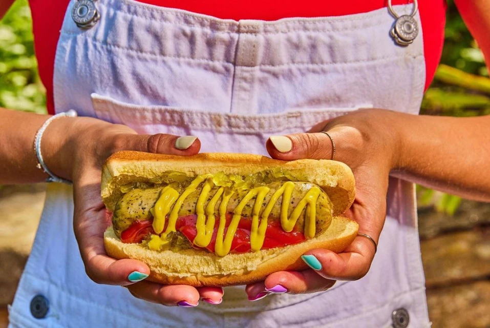 Close-up of a person holding a hot dog with mustard, relish, and tomato in a white bun, outdoors on a sunny day.