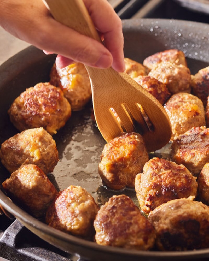 Pan of golden-brown meatballs being cooked on a stovetop and stirred with a wooden cooking spoon for a homemade meal.