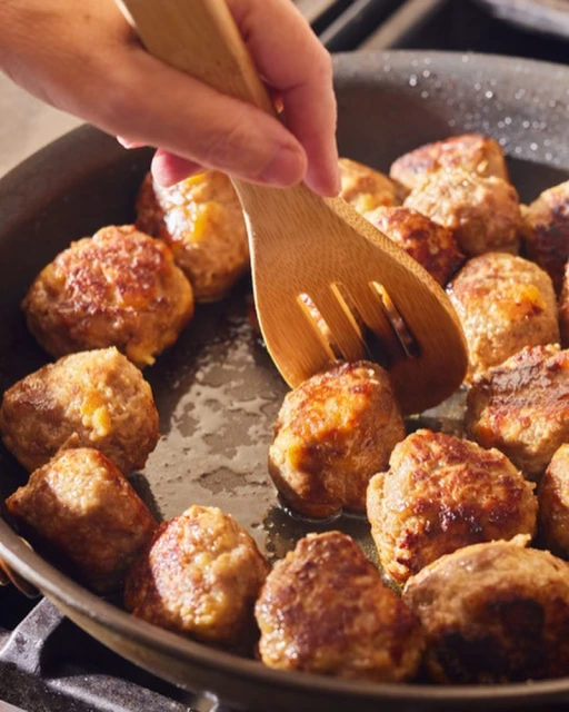 Pan of golden-brown meatballs being cooked on a stovetop and stirred with a wooden cooking spoon for a homemade meal.