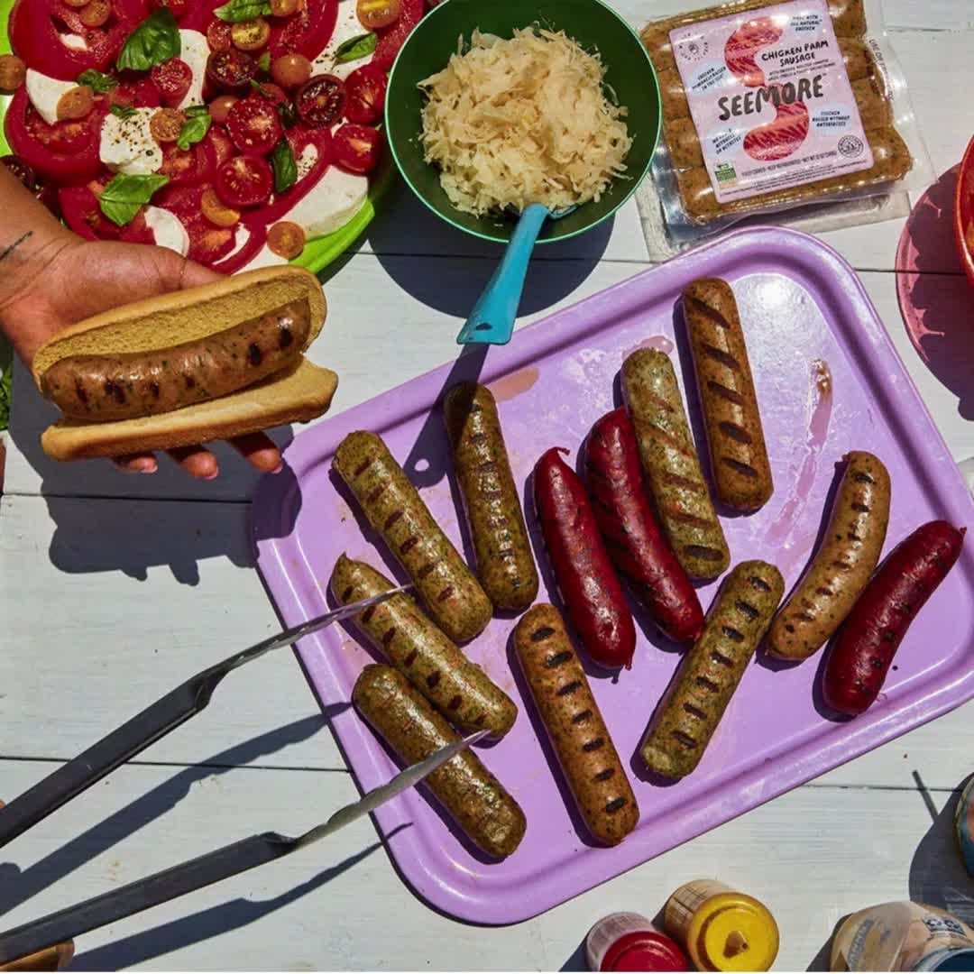 Overhead view of grilled sausages on a purple tray, a sausage in a bun in a hand, tomato salad, and sauerkraut complete a tasty meal.