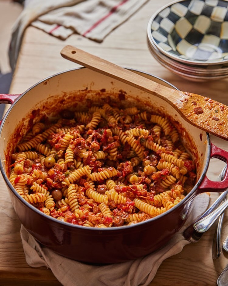 Overhead shot of red enameled cast iron pot filled with spiral pasta in tomato sauce with wooden spoon, plates, and silverware.