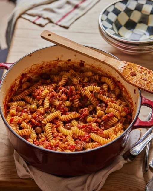 Overhead shot of red enameled cast iron pot filled with spiral pasta in tomato sauce with wooden spoon, plates, and silverware.