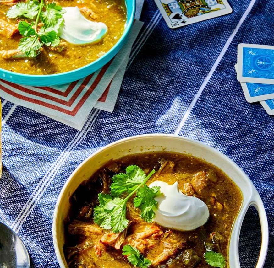 Two bowls of green chili with sour cream and cilantro on a blue checkered tablecloth, along with playing cards, in a bright, natural light.