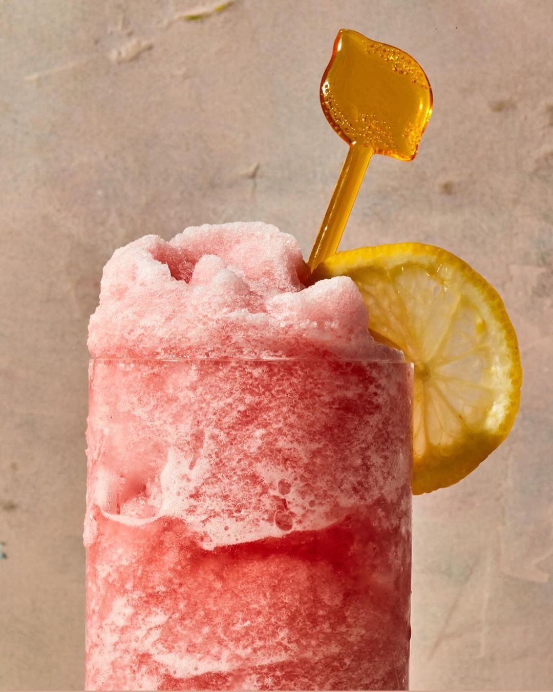 Frosted pink beverage in a glass with lemon slice and yellow straw, illuminated by soft lighting against a textured backdrop.