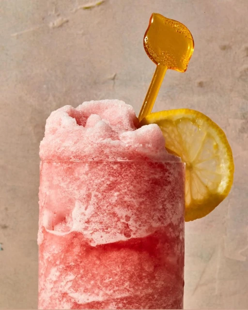Frosted pink beverage in a glass with lemon slice and yellow straw, illuminated by soft lighting against a textured backdrop.