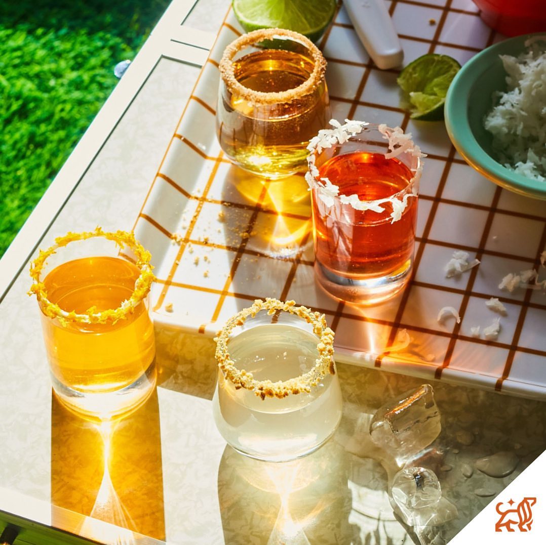 Close-up of four shot glasses with rim decorations and colorful liquids on a reflective surface with grass in the background.