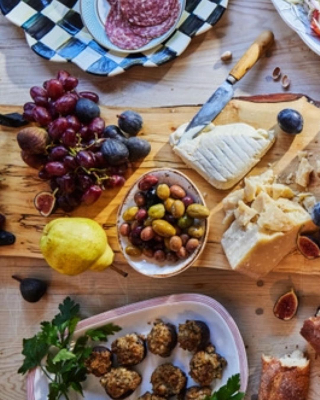 Overhead shot of a wooden board laden with grapes, figs, olives, cheeses, and a pear, complemented by plates of appetizers and charcuterie.