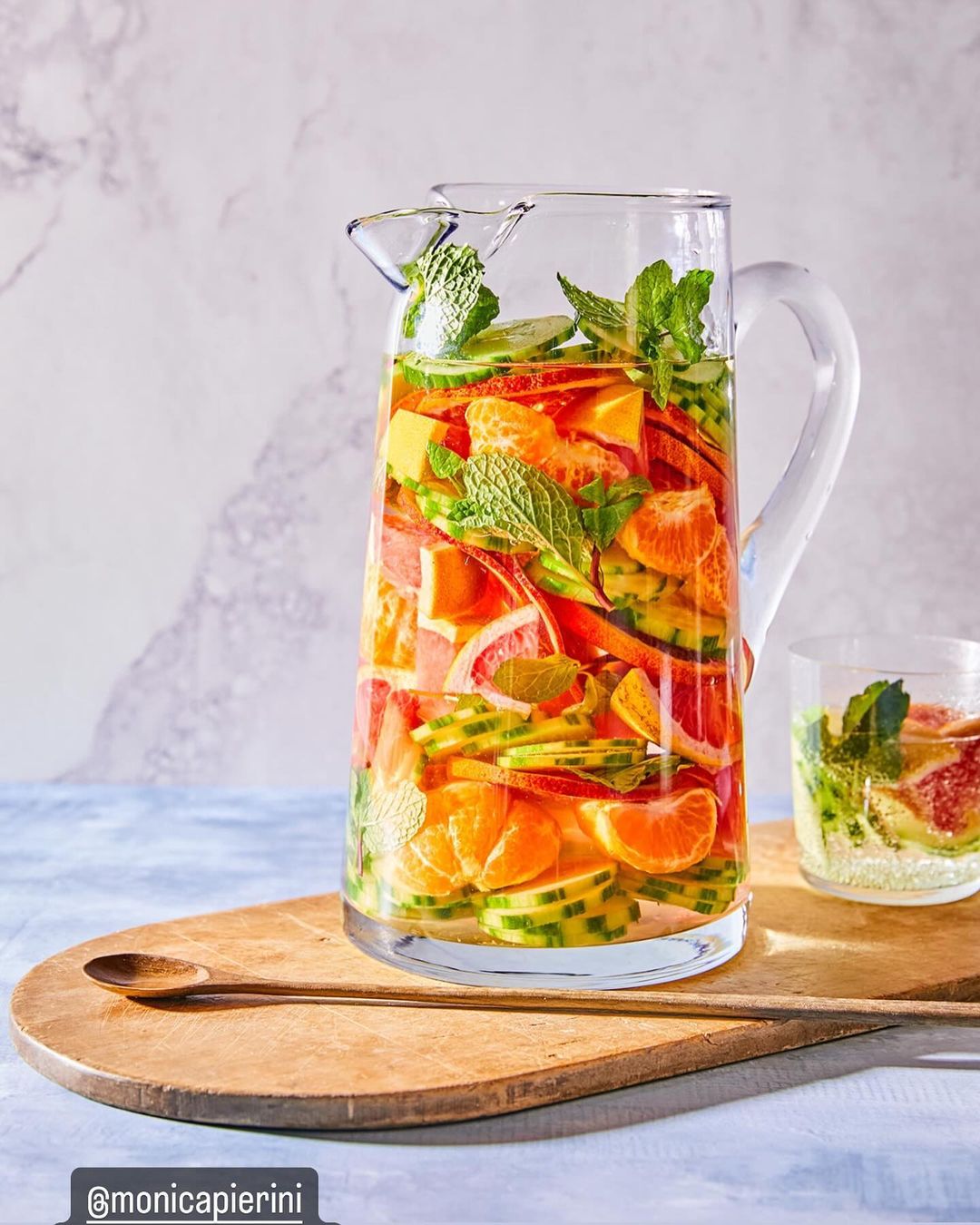 Clear pitcher filled with fruit-infused water including oranges, grapefruit, cucumbers, and mint leaves on a wooden serving board.