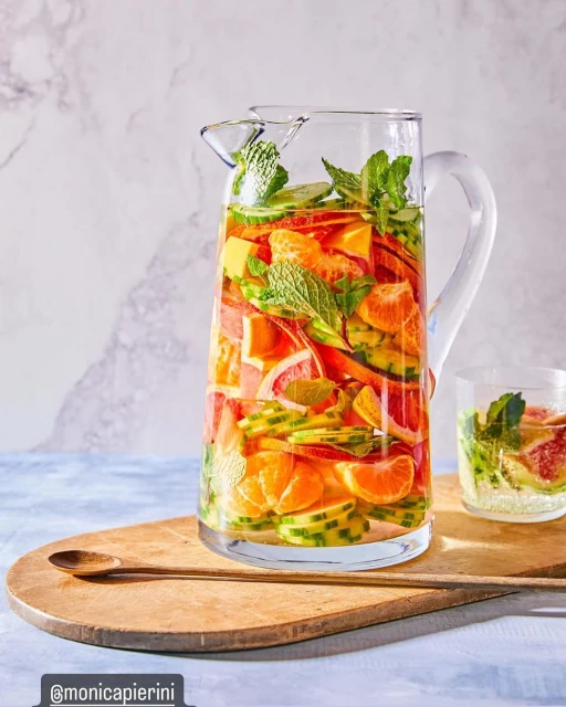 Clear pitcher filled with fruit-infused water including oranges, grapefruit, cucumbers, and mint leaves on a wooden serving board.