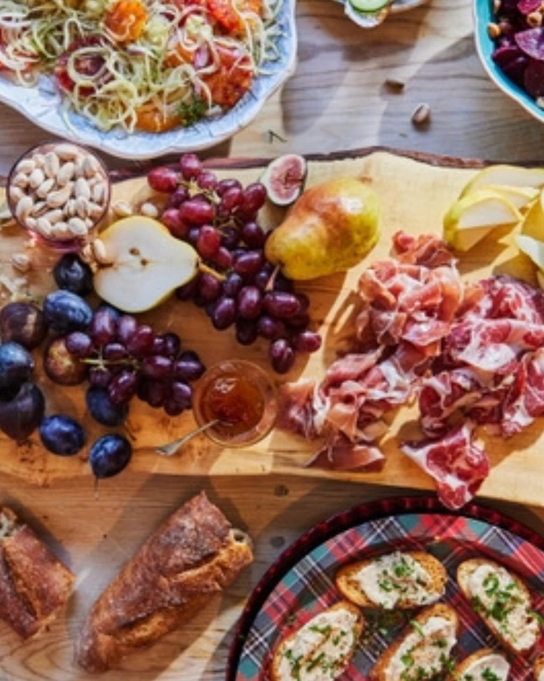 Overhead shot of a wooden charcuterie board featuring a variety of food including grapes, sliced meats, pears, bread and salad.