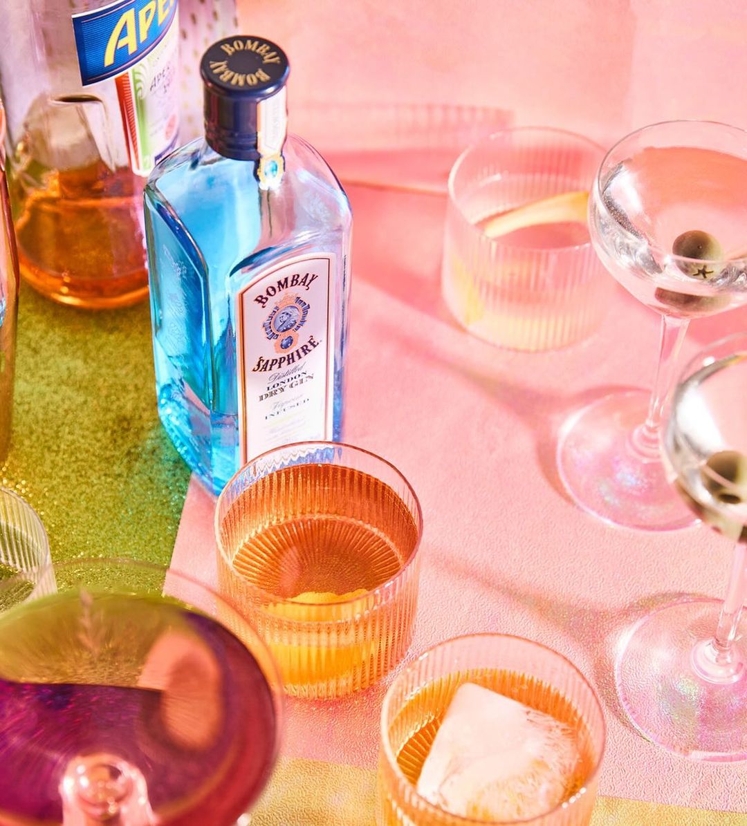Overhead shot of cocktail glasses and bottles on a pink and green reflective surface with pink lighting, capturing a vibrant and stylish bar scene.