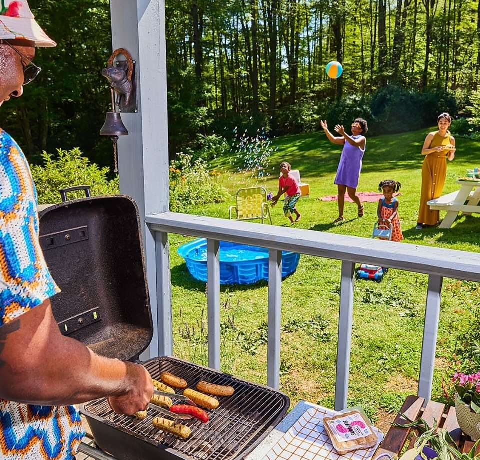 Family enjoying a summer cookout in their backyard, with an adult cooking on a grill, kids playing, and a swimming pool visible in the background.