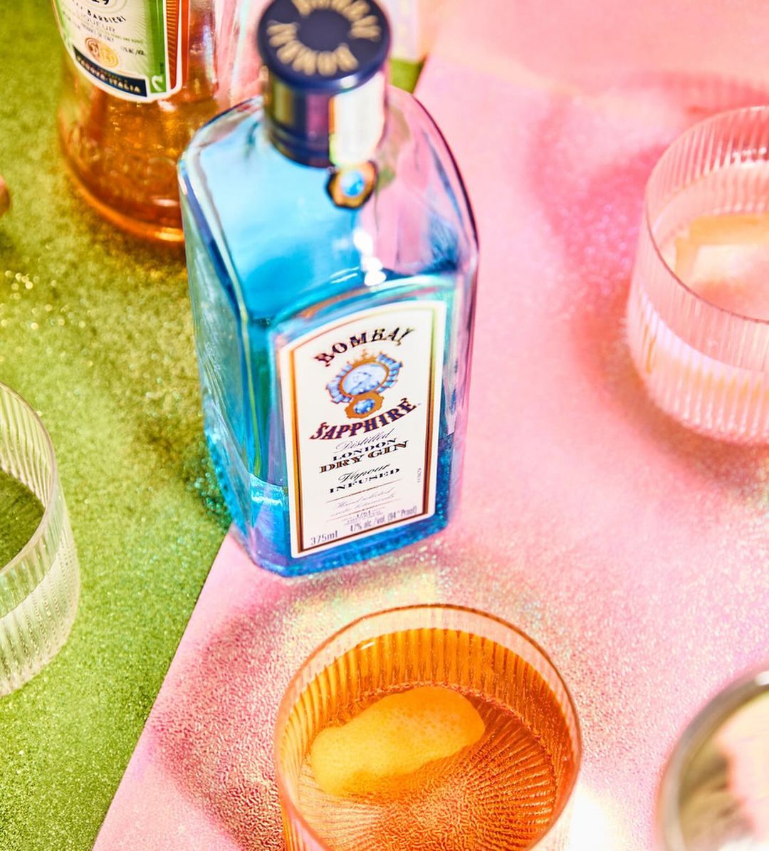 Bombay Sapphire gin bottle and cocktail glasses with drinks on a glittery pink and green surface, studio shot for spirits advertising.