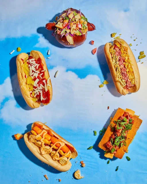 Overhead shot of an assortment of unique hot dogs with diverse toppings arranged in a circle against a light blue cloudy background.