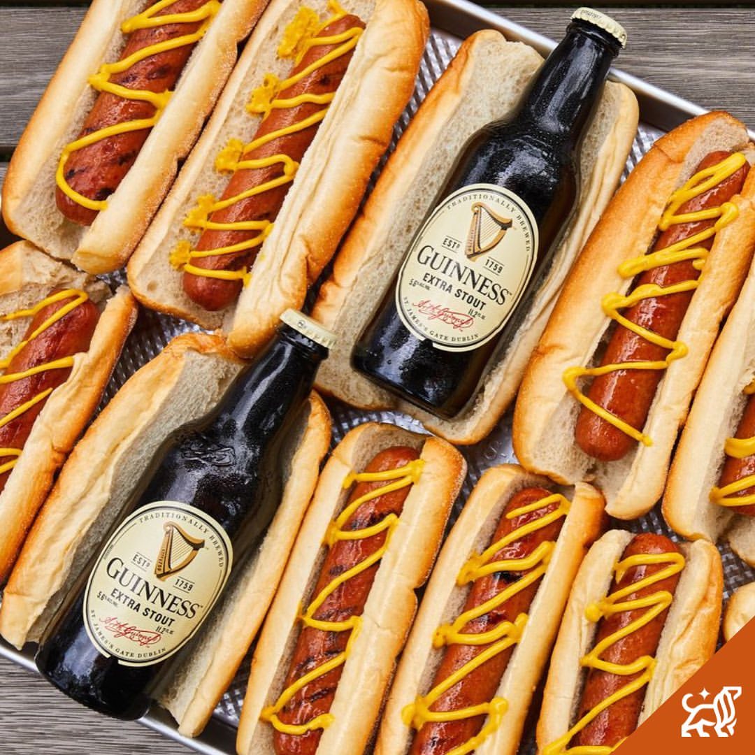 Overhead shot of hot dogs with mustard and two bottles of Guinness Extra Stout beer on a metal serving tray.