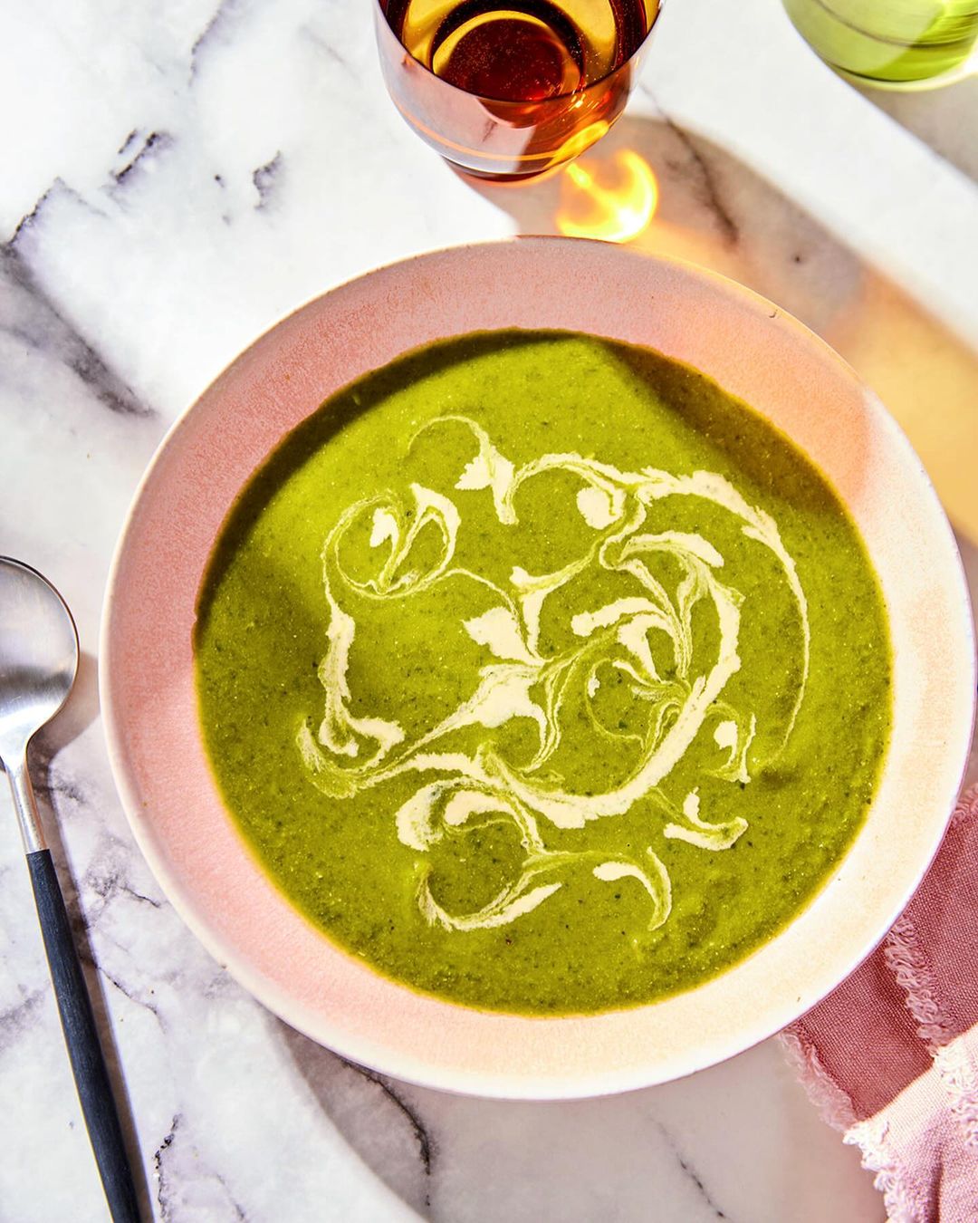 Overhead shot of a bowl of green soup with cream swirl on a marble surface, alongside a spoon, linen, and drinkware, with soft lighting.