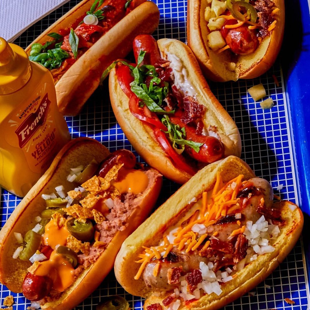 Assorted hot dogs with various toppings like tomatoes, onions, peppers, jalapenos, bacon, and mustard on a blue grid surface.