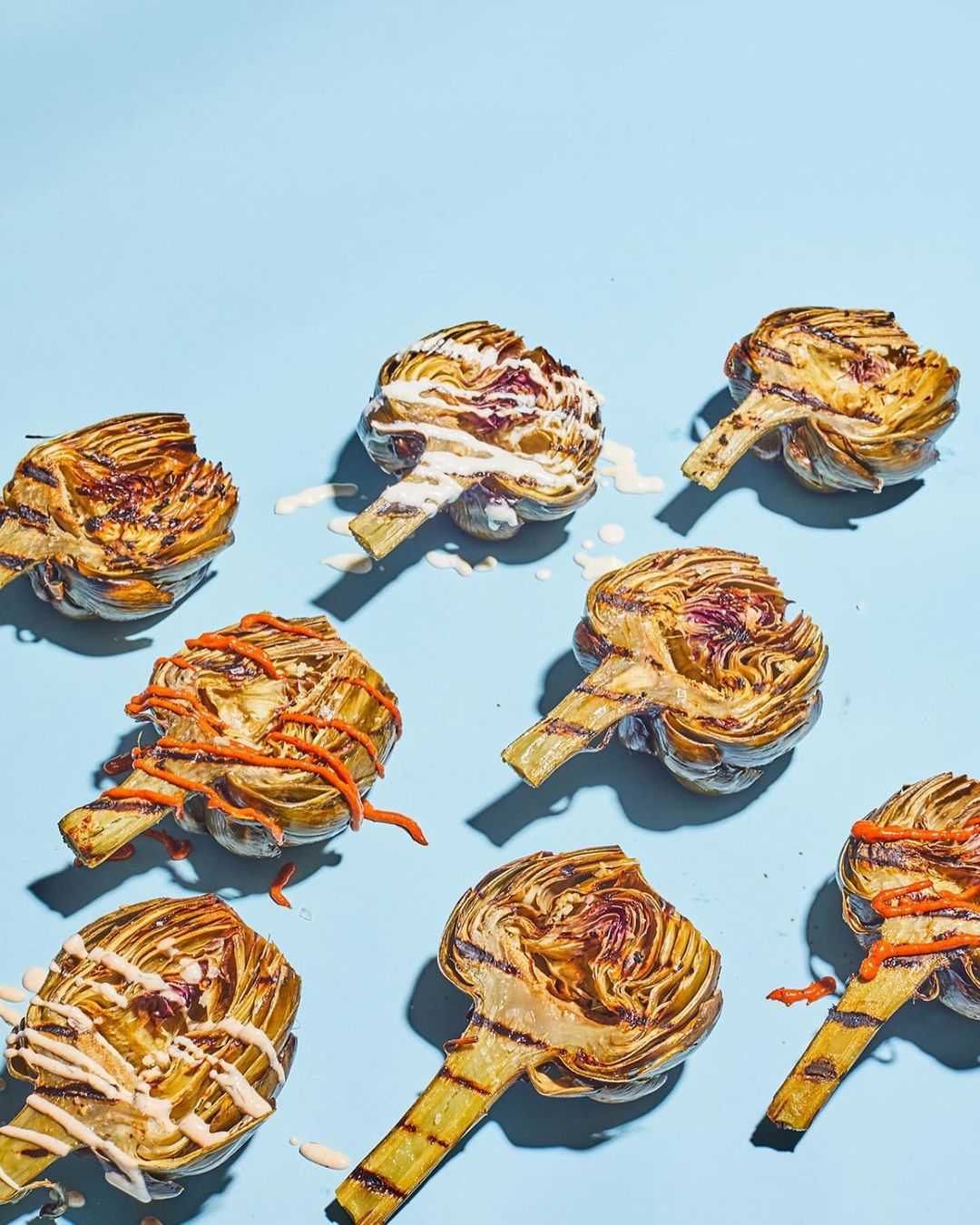 Grilled artichokes with white and orange sauces are arranged on a light blue background in a studio food photography shot.