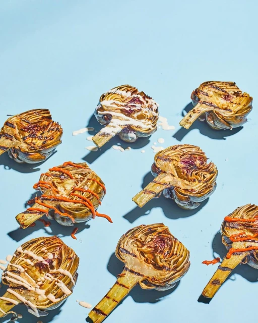 Grilled artichokes with white and orange sauces are arranged on a light blue background in a studio food photography shot.