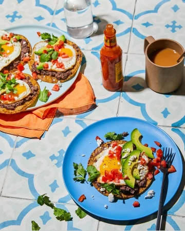Overhead shot of tostadas with fried eggs, avocado, tomatoes and cilantro, served on blue plates with hot sauce and coffee.