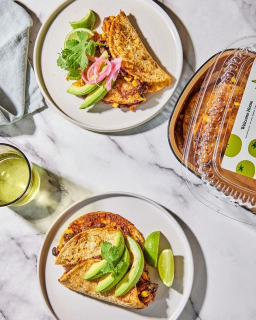 Two plates feature stacked quesadillas, garnished with avocado slices, lime wedges, and cilantro, alongside a glass of white liquid.