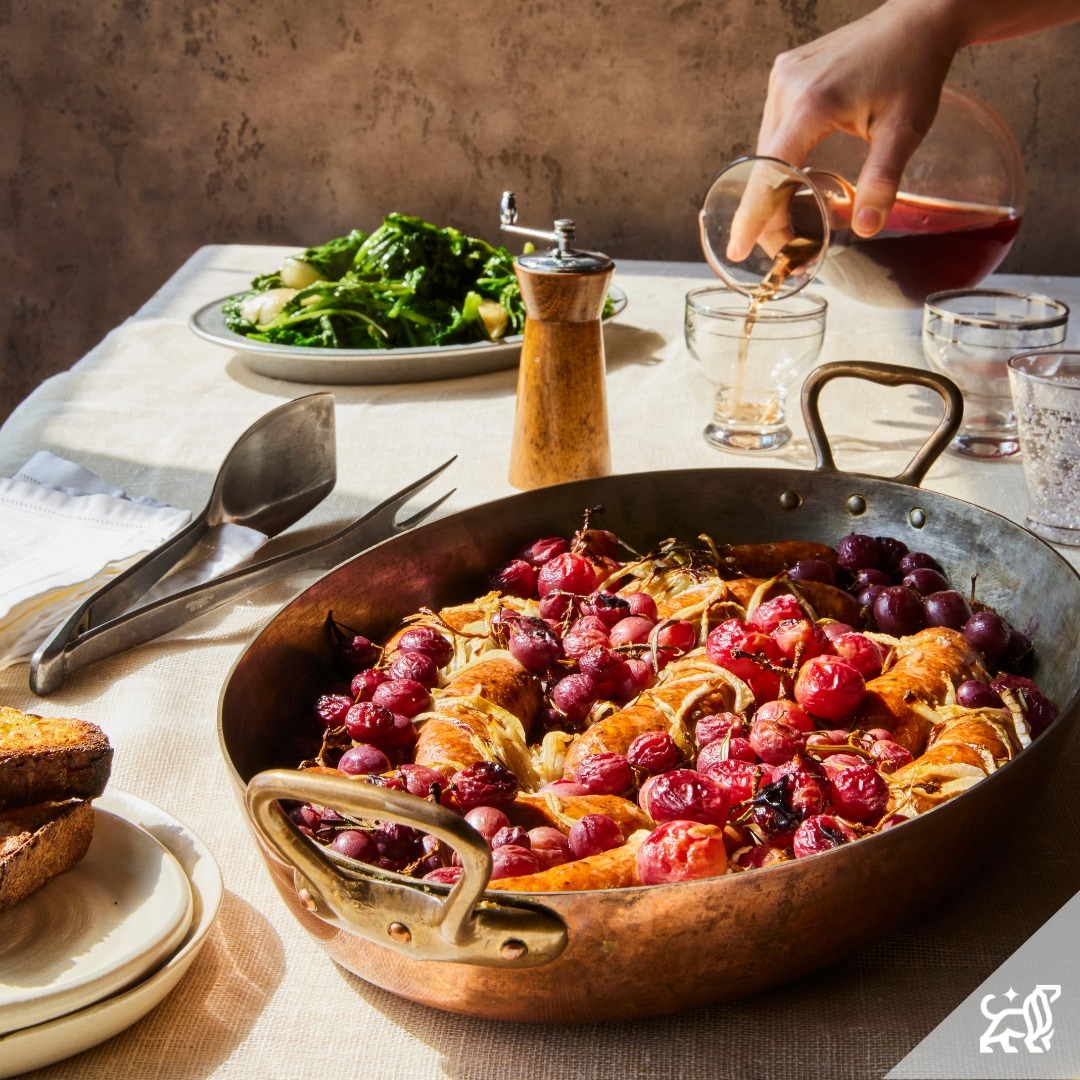Overhead shot of sausage and grapes dish being prepared, wine being poured on top, spinach salad, and a pepper grinder in the background.