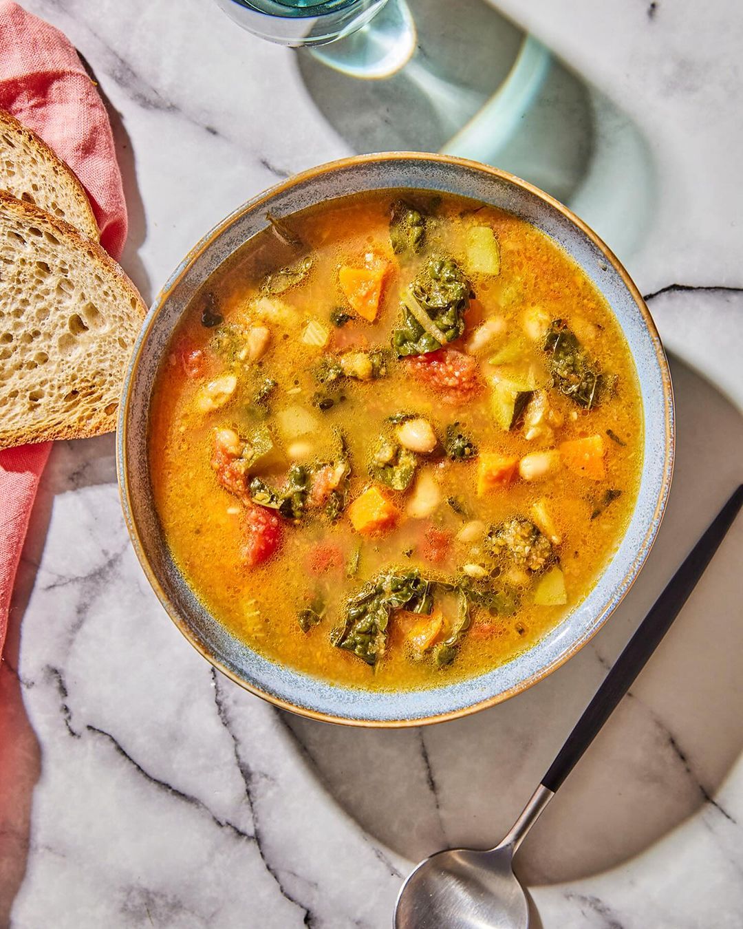 Overhead shot of a bowl of vegetable soup with kale, white beans, tomatoes, and squash, served with a spoon and slices of bread.
