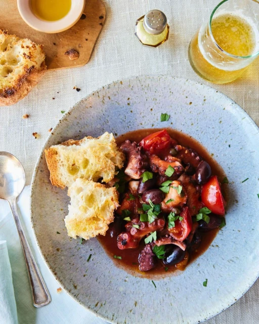 Overhead shot of octopus stew with tomatoes and olives in a speckled ceramic bowl, served with grilled bread and a beverage.