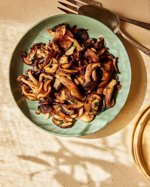 Overhead shot of cooked mushrooms with herbs on a green plate, paired with silverware, on a tan surface with soft lighting.