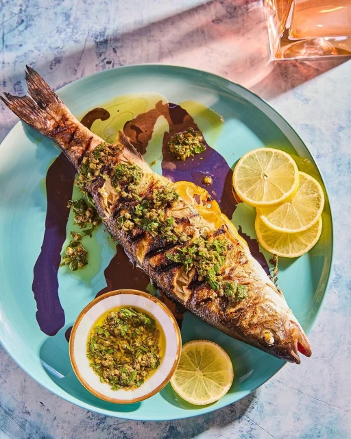 Grilled fish plated with lemon slices and green sauce. Food presentation featuring seafood on a turquoise plate with colorful backdrop.