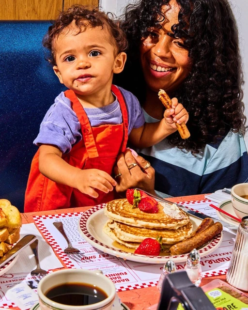 Toddler with mother sitting at a table in a diner with pancakes, sausage, coffee, and toast, family enjoying brunch in a restaurant.