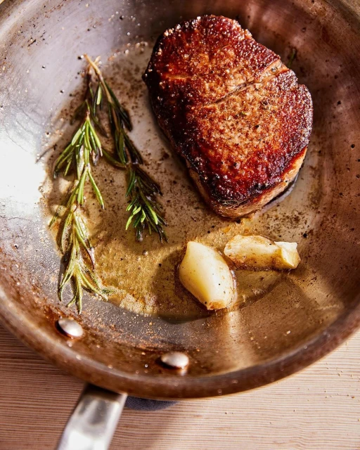 Pan-seared steak with garlic and rosemary in a silver skillet, showcasing a culinary delight with savory herbs and aromatics.