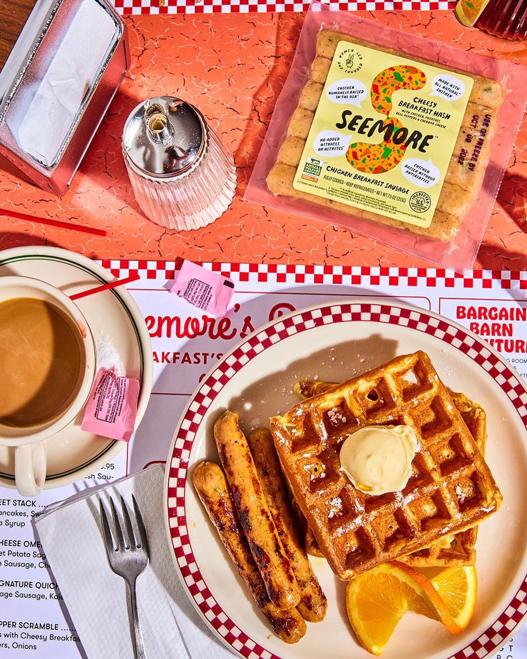 Breakfast plate featuring waffles with butter, sausages, and orange slices accompanied by coffee on a red-and-white checkered table.