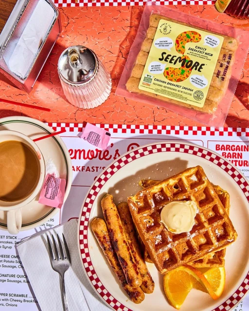 Breakfast plate featuring waffles with butter, sausages, and orange slices accompanied by coffee on a red-and-white checkered table.