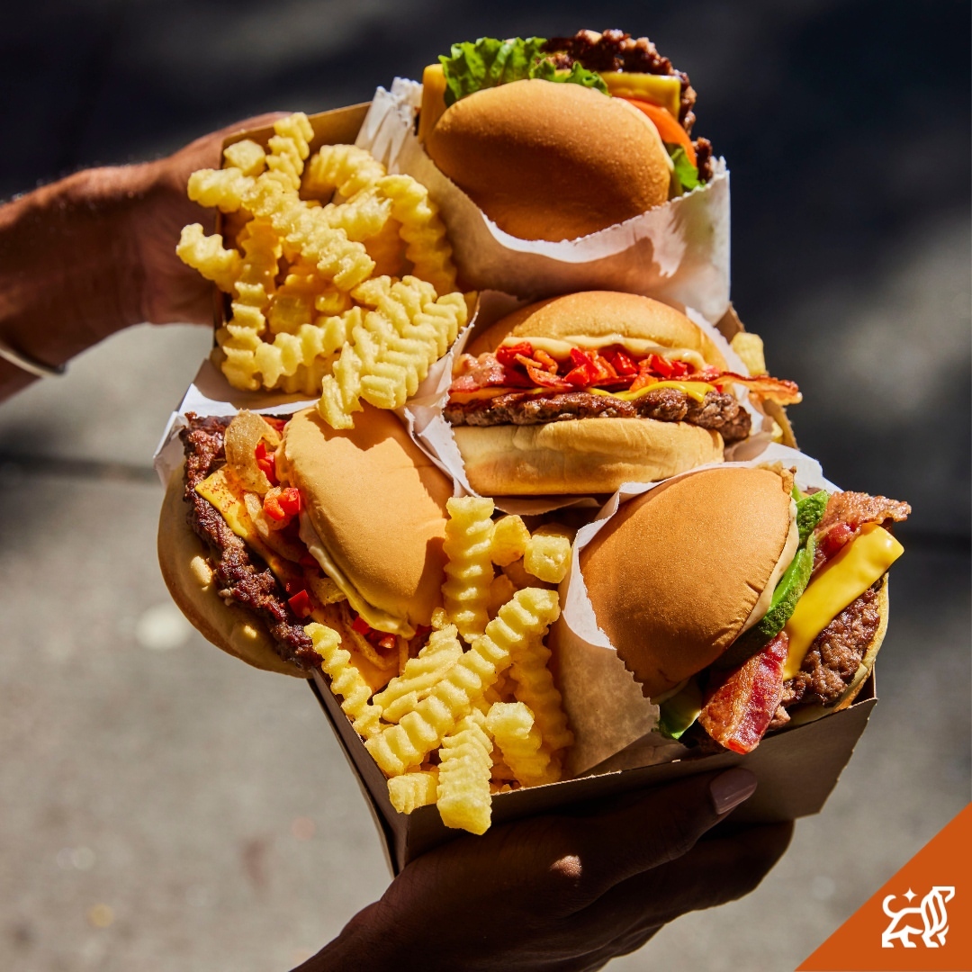 Four burgers and fries are held in hands, showcasing stacked sandwiches with various toppings, cheddar cheese, bacon, and fresh lettuce.