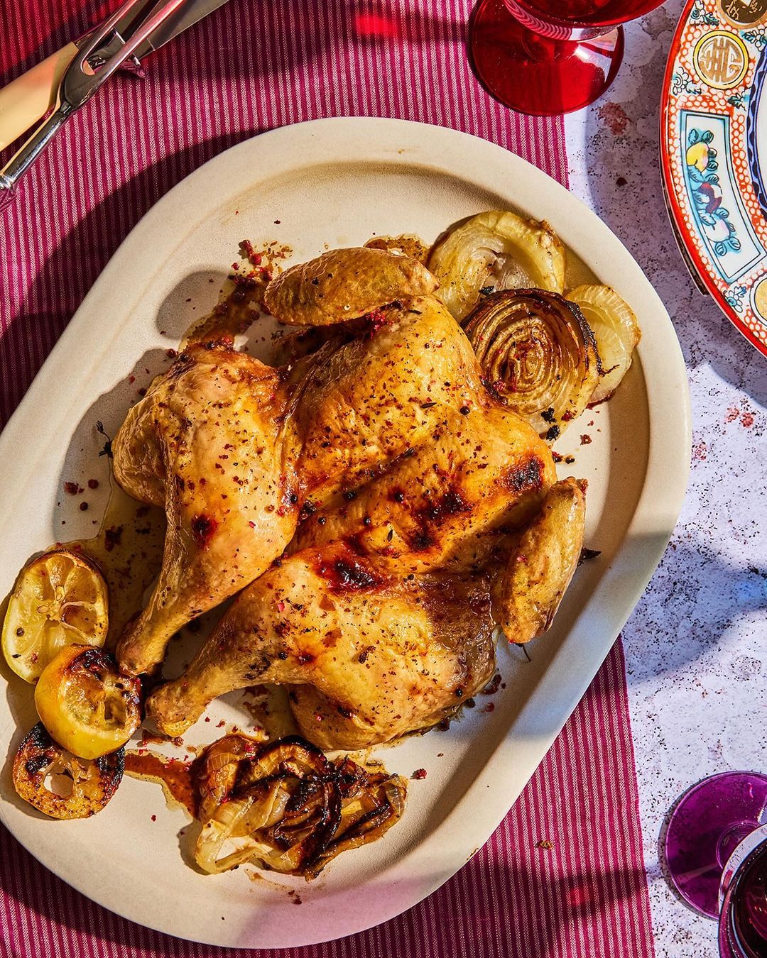 Overhead shot of a golden-brown roasted chicken served on an oval platter with caramelized onions and grilled lemon halves, with pink tablecloth.