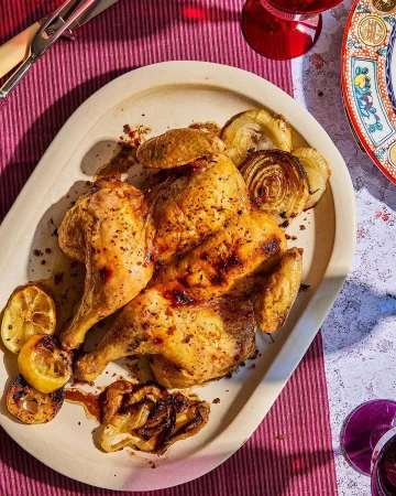 Overhead shot of a golden-brown roasted chicken served on an oval platter with caramelized onions and grilled lemon halves, with pink tablecloth.