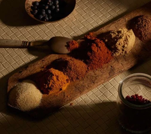 Overhead shot of wooden board with piles of colorful spices like turmeric, paprika, and pepper, with spoon, bowl, and jar.