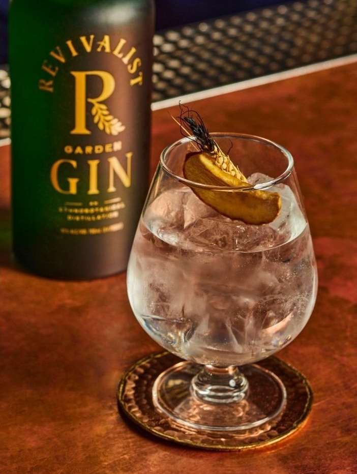 Gin cocktail garnished with citrus peel, served in a stemmed glass on a copper coaster with a bottle of gin in the background.