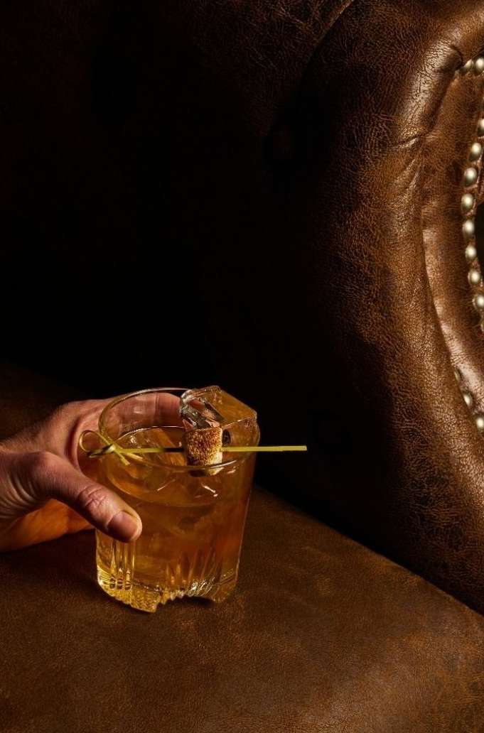 A hand holds a crystal glass of amber cocktail on a leather surface next to a leather chair, showcasing an alcoholic beverage.
