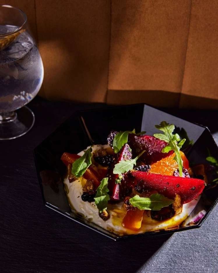 Close-up shot of a salad on a black plate next to a water glass, featuring beets, oranges, greens, and a creamy sauce, on a dark surface.