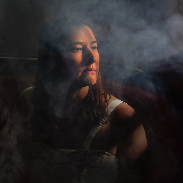 Adult female portrait, eye-level view, ethereal smoke, dark background. Dressed in a white top, she gazes left in a semi-lit studio.