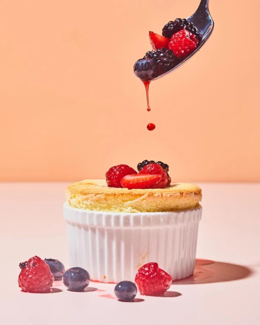 Close-up of a white souffle ramekin topped with fresh raspberries, strawberries, and blueberries and a spoon with berries dripping.