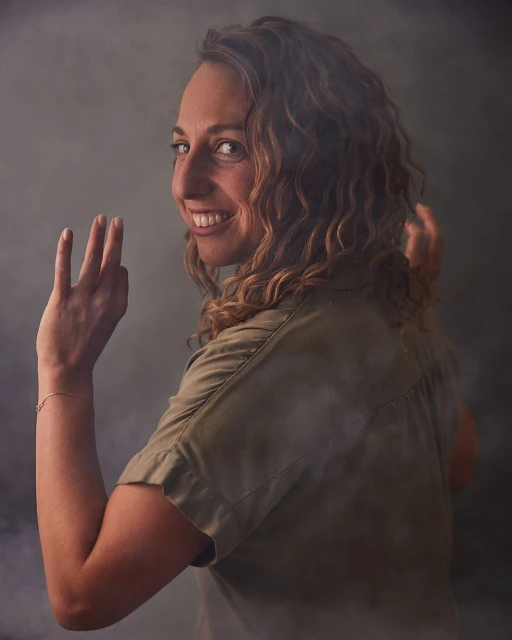 Smiling woman with curly hair looks over her shoulder, raising her hand in a playful gesture against a smokey grey background.  