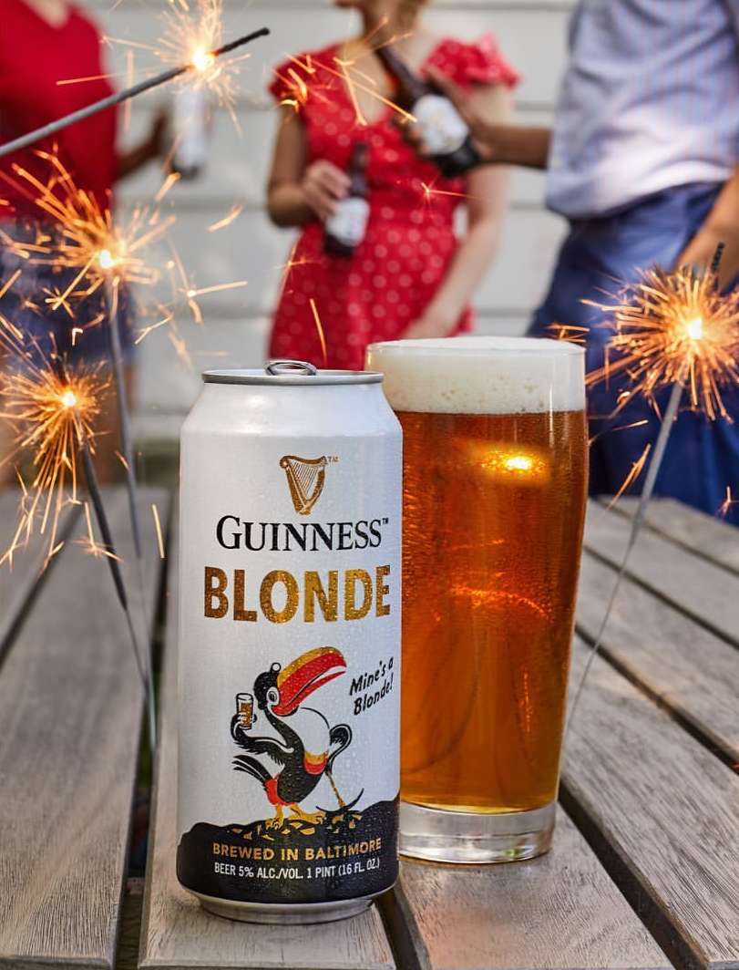 Close-up of Guinness Blonde beer can and full pint glass on wood surface, with sparklers and group of people in background.