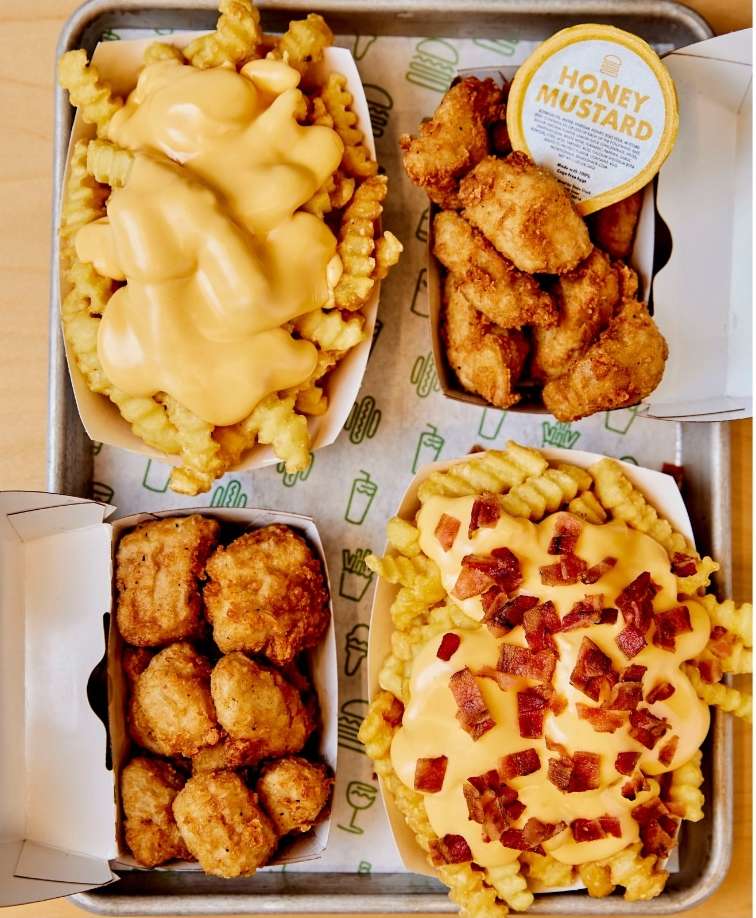 Overhead shot of fries with cheese and bacon, chicken nuggets, and honey mustard on a gray tray. Fast-food concept.