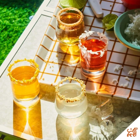 Four colorful cocktail shots with salted rims on a square tiled tray with limes and shredded coconut