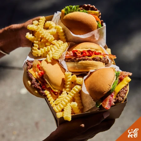 Close-up of a person holding a cardboard carton filled with four gourmet cheeseburgers and crinkle-cut french fries.