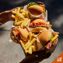 Close-up of a person holding a cardboard carton filled with four gourmet cheeseburgers and crinkle-cut french fries.